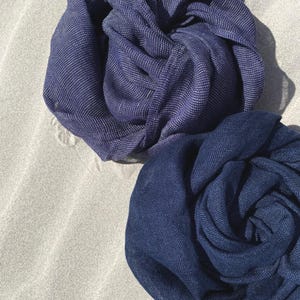 May include: Two navy blue scarves, artfully arranged, with a textured appearance. One scarf has a subtle silver thread woven throughout, adding a touch of shimmer. The scarves are displayed on a neutral, sandy surface.