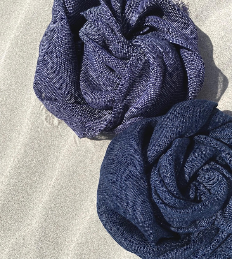 May include: Two navy blue scarves, artfully arranged, with a textured appearance. One scarf has a subtle silver thread woven throughout, adding a touch of shimmer. The scarves are displayed on a neutral, sandy surface.