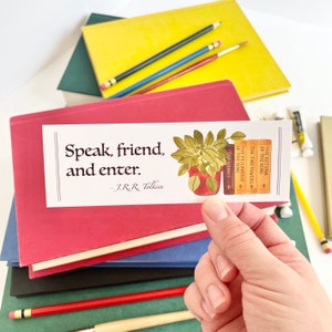 May include: A rectangular bookmark with the quote "Speak, friend, and enter" by J.R.R. Tolkien. The bookmark features a watercolor illustration of a potted plant and a stack of books. The books are titled "The Hobbit", "The Fellowship of the Ring", "The Two Towers", and "The Return of the King".