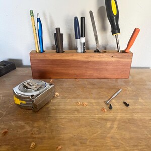 May include: A wooden organizer with six slots for holding tools and writing utensils. The organizer is made of reddish-brown wood and has a natural finish. The slots are filled with a variety of items, including a pencil, pens, markers, a screwdriver, a ruler, and a small orange tool.