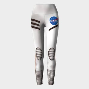 May include: White leggings featuring a NASA logo on the upper left thigh. The design includes dark horizontal stripes on the right hip, ribbed knee patches, and horizontal line patterns at the ankles, mimicking the appearance of a space suit.