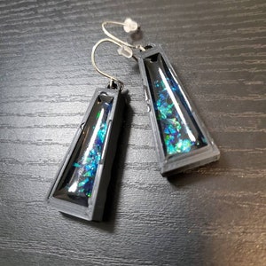 May include: A pair of black triangle-shaped earrings with a clear resin insert filled with iridescent blue and green glitter.