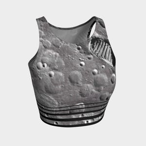 May include: A sleeveless crop top featuring a high-resolution photographic print of the lunar surface, complete with craters and a prominent boot print. The garment includes a series of horizontal black stripes along the bottom hem.