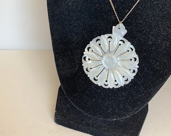 Puede incluir: Un collar con un colgante de nácar blanco, adornado con un delicado diseño floral. El colgante es redondo y presenta una flor tallada con una piedra central. El collar está en una cadena de plata.
