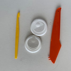 May include: Two white plastic lids with a small white circle in the center, a yellow plastic scraper, and an orange plastic knife with the text "JUBILEE WARE" printed on the side.