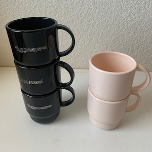 May include: A stack of four black mugs with the word "Tupperware" printed on them, and a stack of three pink mugs. All mugs have handles.