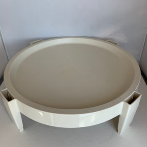 May include: White plastic round cake stand with four legs. The stand is approximately 12 inches in diameter.