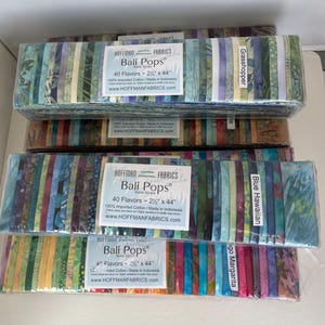 May include: Three stacks of Hoffman Fabrics Bali Pops batik strips in plastic packaging. Each stack contains 40 fabric strips measuring 2.5 inches by 44 inches. The top stack is labeled Grasshopper, the middle Blue Hawaiian, and the bottom Mango Margarita.