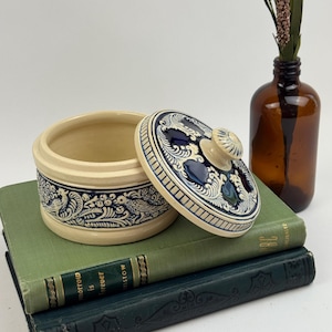 May include: A round ceramic jar with a removable lid, featuring a cream-colored base and deep blue embossed patterns of birds and foliage. The jar is displayed alongside a brown glass bottle and two stacked books with green covers.