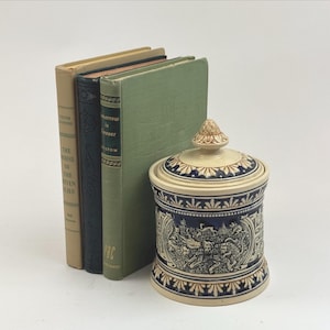 May include: A stack of three hardcover books in tan, black, and green, positioned next to a cylindrical ceramic jar with a lid. The jar features a deep blue and cream color scheme with intricate embossed relief patterns of figures and foliage.