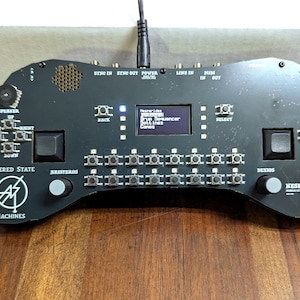 Puede incluir: Dispositivo negro de producción de música electrónica con múltiples botones, perillas y una pequeña pantalla que muestra texto. El dispositivo tiene el texto "ALTERED STATE MACHINES" y "HESPERIDES" impreso. Un cable de alimentación está enchufado en la parte superior.