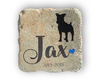 May include: A square, light-colored stone with a black silhouette of a dog and the name "Jax" in a decorative font. A blue heart is next to the name, and the years "2015-2018" are below.