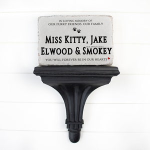 May include: A rectangular light gray stone memorial plaque with rough edges resting on a black wall shelf. The text reads: IN LOVING MEMORY OF OUR FURRY FRIENDS. OUR FAMILY, MISS KITTY, JAKE, OUR SWEET LITTLE ELWOOD & SMOKEY, YOU WILL FOREVER BE IN OUR HEARTS.
