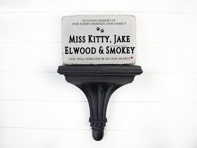 May include: A rectangular light gray stone memorial plaque with rough edges resting on a black wall shelf. The text reads: IN LOVING MEMORY OF OUR FURRY FRIENDS. OUR FAMILY, MISS KITTY, JAKE, OUR SWEET LITTLE ELWOOD & SMOKEY, YOU WILL FOREVER BE IN OUR HEARTS.