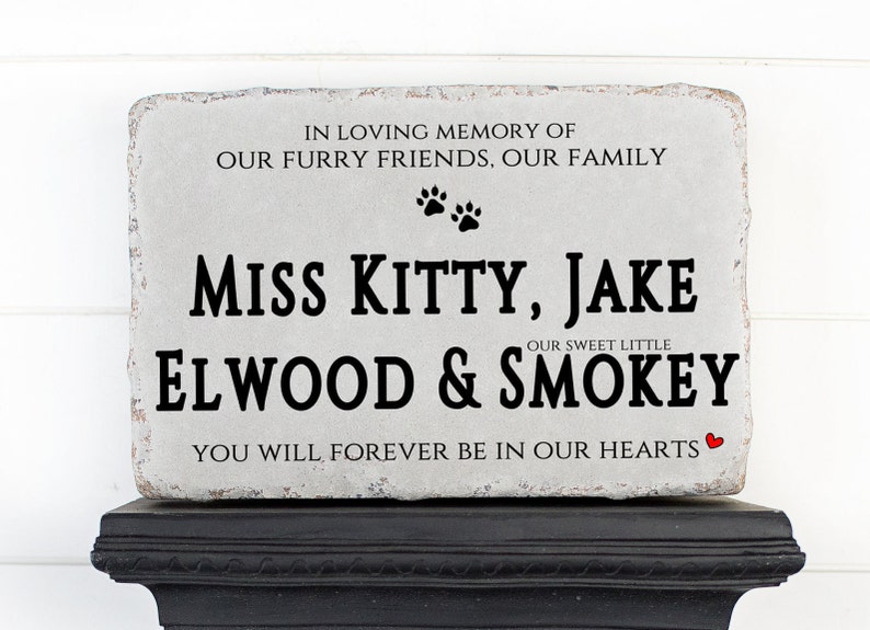 May include: A rectangular gray stone memorial plaque with rough edges featuring black text that reads IN LOVING MEMORY OF OUR FURRY FRIENDS, OUR FAMILY, MISS KITTY, JAKE, OUR SWEET LITTLE ELWOOD & SMOKEY, YOU WILL FOREVER BE IN OUR HEARTS with two paw prints and a small red heart.