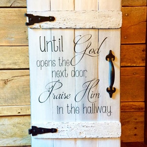 May include: A white wooden sign with the text "Until God opens the next door, Praise Him in the hallway." The sign has a distressed finish, black hinges, and a black handle. The background is a wooden wall.