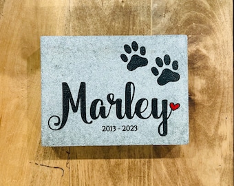 May include: A rectangular gray stone memorial plaque featuring the name Marley in black script, two black paw prints, a small red heart, and the years 2013-2023 printed in black. The surface has a speckled texture and a flat, block-like shape.