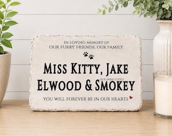 May include: A rectangular stone memorial plaque with rough, textured edges. The text reads: IN LOVING MEMORY OF OUR FURRY FRIENDS, OUR FAMILY, MISS KITTY, JAKE, OUR SWEET LITTLE ELWOOD & SMOKEY, YOU WILL FOREVER BE IN OUR HEARTS, with two paw prints.