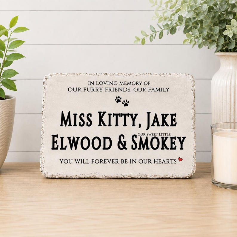 May include: A rectangular stone memorial plaque with rough, textured edges. The text reads: IN LOVING MEMORY OF OUR FURRY FRIENDS, OUR FAMILY, MISS KITTY, JAKE, OUR SWEET LITTLE ELWOOD & SMOKEY, YOU WILL FOREVER BE IN OUR HEARTS, with two paw prints.