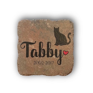 May include: A square, weathered stone with the name "Tabby" in black script, a black cat silhouette, and the years 2002-2017. A small red heart is next to the name. The stone has a natural, earthy color.