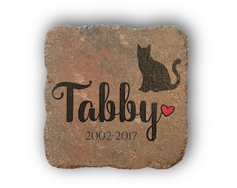 May include: A square, weathered stone with the name "Tabby" in black script, a black cat silhouette, and the years 2002-2017. A small red heart is next to the name. The stone has a natural, earthy color.