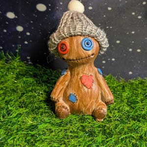 May include: A handcrafted brown figurine with a stitched heart and patches. It has red and blue eyes and wears a gray knit cap with a white pom-pom. The figurine is posed on green grass against a starry background.