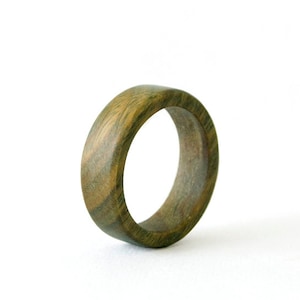Può includere: Un anello in legno di colore verde oliva naturale. L'anello è liscio e ha una forma arrotondata. La venatura del legno è visibile, aggiungendo consistenza. Diametro di circa 2,5 cm.