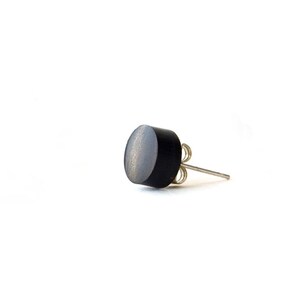 May include: A single stud earring featuring a circular, matte black face with a visible wood grain texture. The earring includes a silver-toned metal post and a matching butterfly clutch backing, designed for secure attachment to an earlobe.
