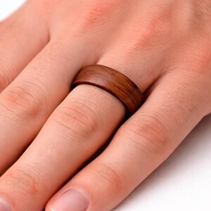 May include: A wooden ring with a rich brown color is worn on a finger. The ring has a smooth, polished surface and a simple, classic design. The ring is approximately 0.3 inches wide.