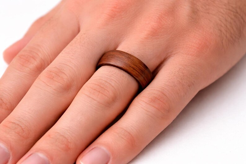 May include: A wooden ring with a rich brown color is worn on a finger. The ring has a smooth, polished surface and a simple, classic design. The ring is approximately 0.3 inches wide.