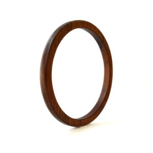 May include: A circular bangle bracelet crafted from dark brown wood with a smooth, polished finish. The bracelet features a simple, continuous ring design with a visible wood grain pattern throughout the surface of the circular wooden band.