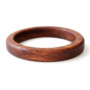 May include: A round, wooden bracelet with a rich, dark brown color. The bracelet is made of a solid piece of wood and has a smooth, polished finish. The bracelet is approximately 2.5 inches in diameter and 0.75 inches wide.