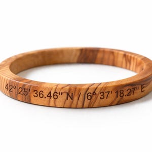 May include: A circular wooden bangle bracelet featuring a natural light brown wood grain pattern. The exterior surface is engraved with the black text 42° 25' 36.46" N / 6° 37' 18.27" E, displaying specific geographic coordinates in a clear, sans-serif font.