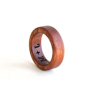 May include: A handcrafted wooden ring with a rich brown hue. The ring features a smooth, polished surface and is engraved with the initials "M + J" on the inside. The ring is a symbol of commitment and love.