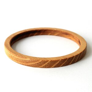 May include: A circular bangle bracelet crafted from light brown wood, featuring a distinct natural grain pattern with diagonal lines running along the outer surface of the band. The wood has a smooth finish and a consistent, thin, rounded profile.
