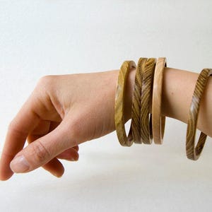 Può includere: Un set di quattro braccialetti rigidi circolari in legno con distinti motivi a venature naturali in tonalità marrone chiaro e medio. I braccialetti sono lisci e levigati, con tre indossati vicini e uno posizionato separatamente sul polso.