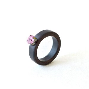 May include: A dark brown wooden ring featuring a single, round, faceted pink gemstone set in a small, dark metal prong setting. The band has a smooth, matte finish and a simple, circular design, highlighting the contrast between the wood and stone.