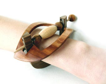 May include: A wooden hair slide featuring a curved, dark brown frame with a central, lighter-toned wooden spindle. The spindle is accented with two small, dark wooden spheres on either side of a smooth, elongated, light-coloured wooden bead.