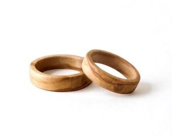May include: Two rings crafted from light brown wood with visible natural grain patterns. The rings feature a smooth, rounded exterior surface and a simple band design, showcasing the organic texture and warm tan tones of the wood material.