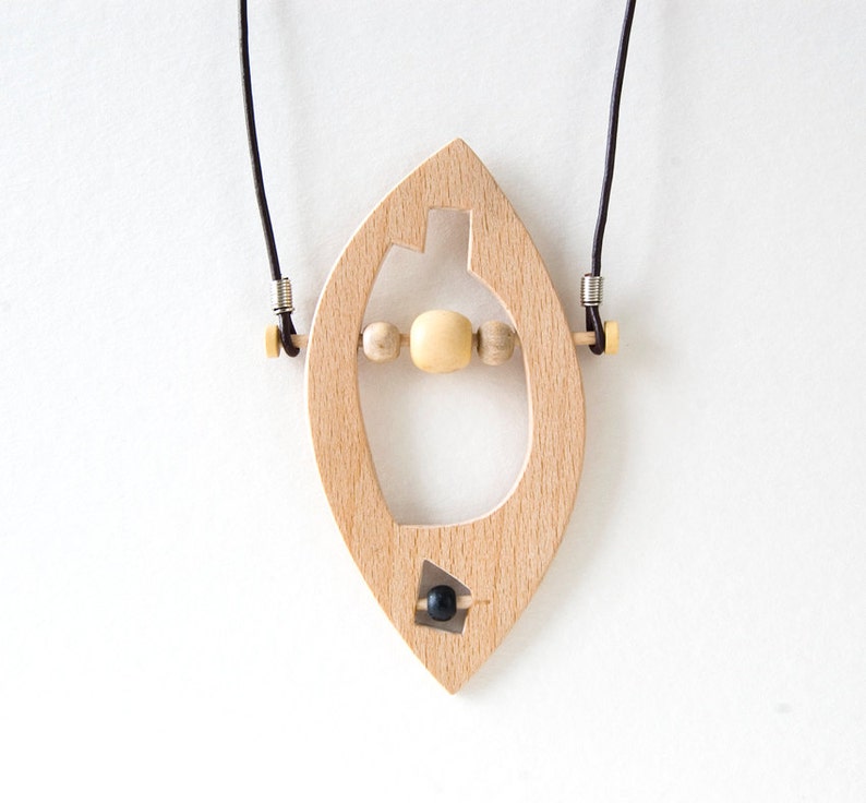 May include: A wooden pendant necklace with a leaf-shaped design. The pendant has a cutout in the shape of a heart and features a small black bead at the bottom. The necklace is made with a brown leather cord and silver metal clasps.