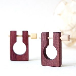 Può includere: Un paio di orecchini in legno con forma quadrata e un ritaglio circolare. Gli orecchini sono in legno rosso scuro e hanno un piccolo piolo in legno che attraversa il centro.