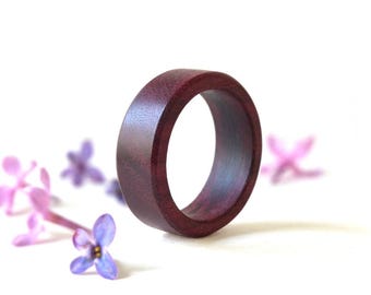 May include: A solid, deep purple-toned wooden ring with a smooth, matte finish and a wide, flat band profile. The ring is positioned upright, showcasing its circular shape and the natural grain texture visible on the dark, rich wood surface.