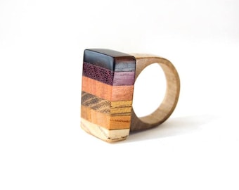 May include: A ring featuring a rectangular face constructed from horizontal layers of wood in varying shades of dark brown, deep purple, reddish-orange, tan, and light cream, attached to a smooth, light-colored wooden band.