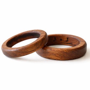 May include: Two wooden rings with a rich brown color. One ring is slightly angled, resting next to the other. The inside of one ring has the engraved initials "J + J". The rings are set against a white background.