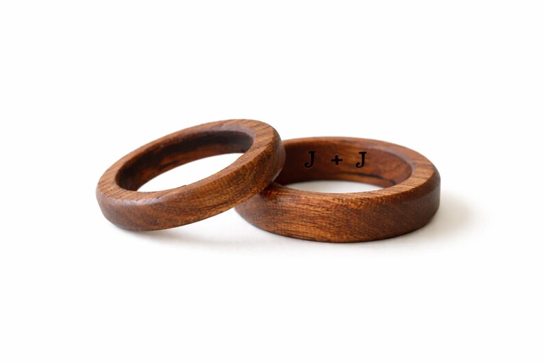 May include: Two wooden rings with a rich brown color. One ring is slightly angled, resting next to the other. The inside of one ring has the engraved initials "J + J". The rings are set against a white background.