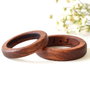 May include: Two wooden rings with a dark brown finish. One ring has the initials "J + J" engraved on the surface. The rings are set against a white background with a hint of greenery.