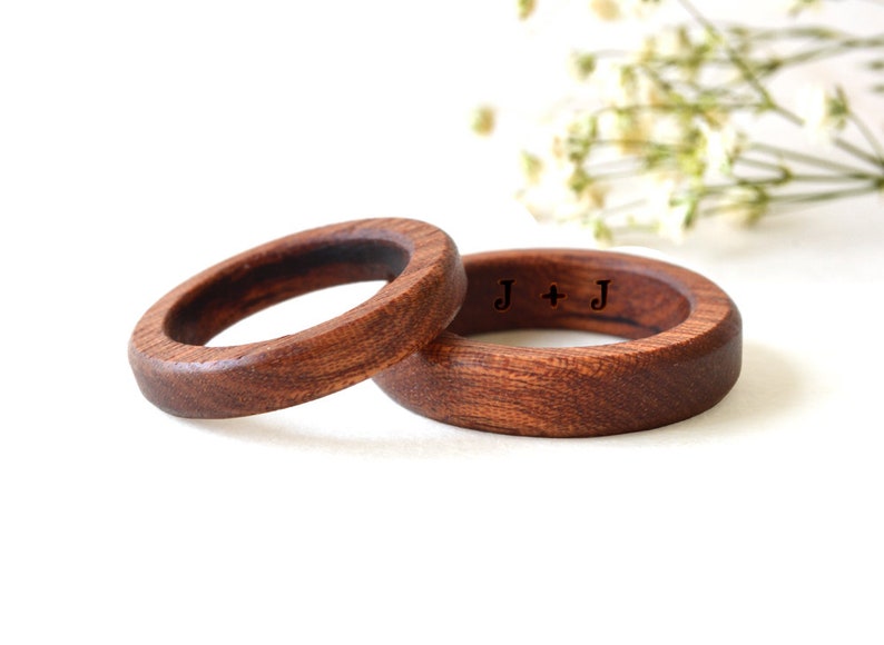 May include: Two wooden rings with a dark brown finish. One ring has the initials "J + J" engraved on the surface. The rings are set against a white background with a hint of greenery.
