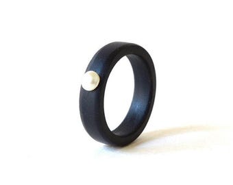 May include: A solid black ring featuring a single, small, round, white pearl set into the outer surface of the band. The ring has a smooth, matte finish and a uniform thickness throughout the circular band, creating a simple, circular jewellery piece.