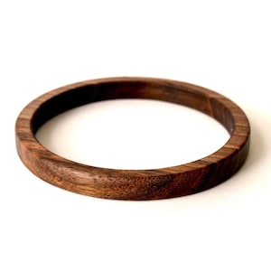 May include: A circular bangle bracelet crafted from dark brown wood with a visible natural grain pattern. The bracelet features a smooth, polished finish and a consistent rounded profile, showcasing the rich, deep tones of the timber material.