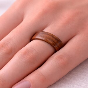 May include: Close-up of a wooden ring with a natural wood grain pattern. The ring is a warm brown color and is worn on a finger. The ring is a simple band, showcasing the wood's natural beauty.
